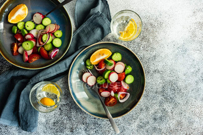 Gläser Zitronenwasser und Schalen mit Gurken, Tomaten, roten Zwiebeln und Rettichsalat — Stockfoto
