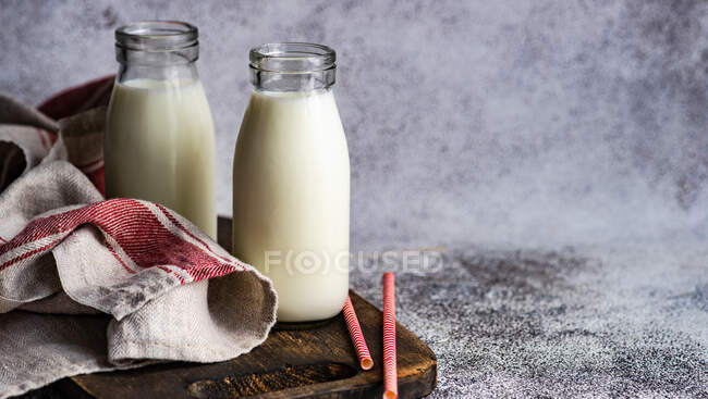 Duas garrafas de leite e palhinhas em uma tábua de corte de madeira com uma toalha de chá — Fotografia de Stock