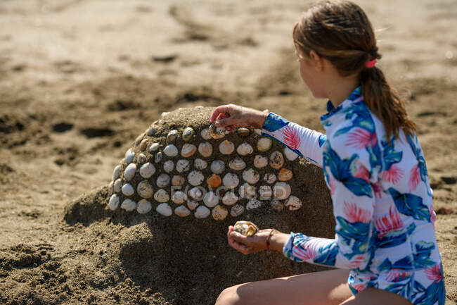 Chica sentada en la playa construyendo una escultura de arena con conchas marinas, Irlanda - foto de stock