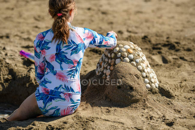 Vista trasera de una niña sentada en la playa construyendo una escultura de arena con conchas marinas, Irlanda - foto de stock