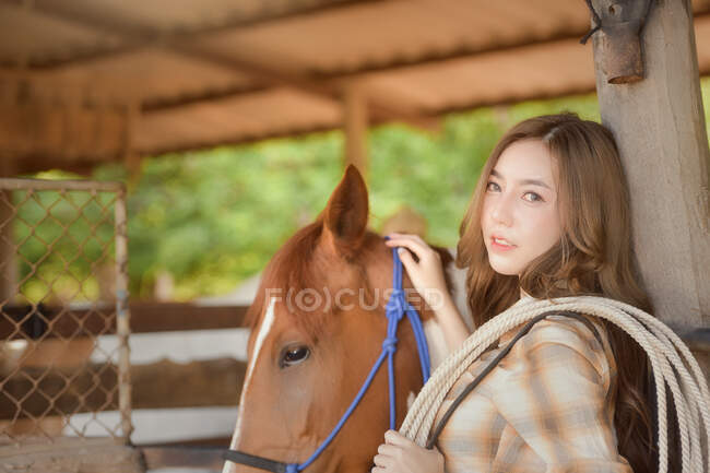 Hermosa mujer de pie en un granero con su caballo, Tailandia - foto de stock