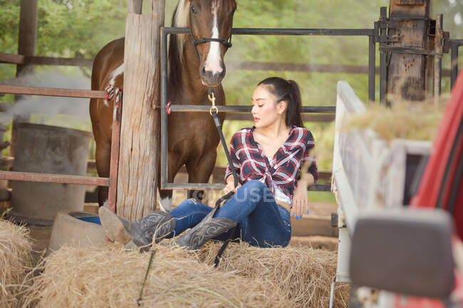 Belle femme assise sur une balle de foin dans une grange avec son cheval, Thaïlande — Photo de stock