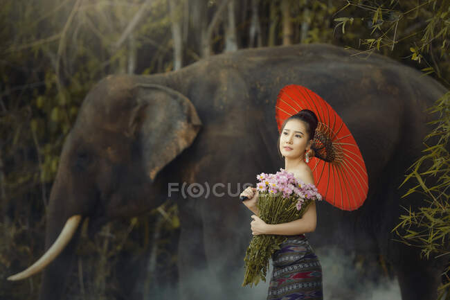 Belle femme tenant un bouquet de fleurs debout près d'un éléphant, Thaïlande — Photo de stock
