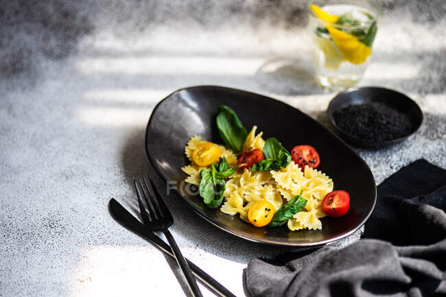 Salad with farfalle pasta and vegetables served in black ceramic bowl with glass of lemon and basil water — Stock Photo