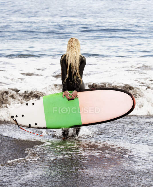 Rear view of a surfer walking into ocean carrying a surfboard behind her back, Bahamas — Stock Photo