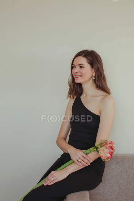 Portrait of a beautiful smiling woman in an evening gown sitting on a sofa holding an Amaryllis flower — Stock Photo