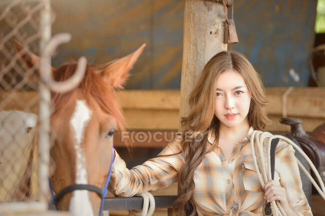 Retrato de una hermosa mujer de pie en un establo con su caballo - foto de stock