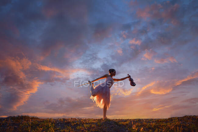 Frau steht auf einem Bein im Freien und hält eine Geige bei Sonnenuntergang, Thailand — Stockfoto