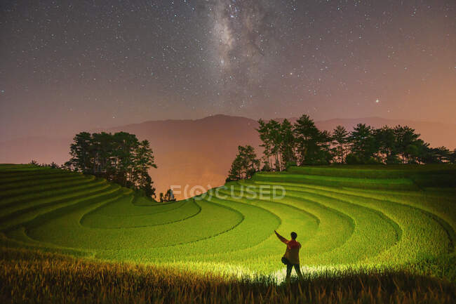 Hombre parado en campos de arroz en terrazas por la noche debajo de la vía láctea, Mu Cang Chai, Yen Bai, Vietnam - foto de stock