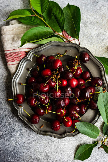 Visão aérea de cerejas frescas em um prato de estanho ao lado de uma toalha de chá e folhagem — Fotografia de Stock