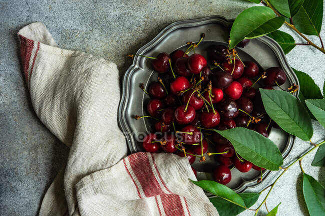 Overhead view of fresh cherries on a pewter plate next to a tea towel and foliage — Stock Photo