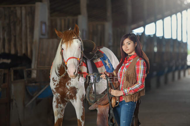 Woman standing in a stable with her horse ready to go riding — Foto stock