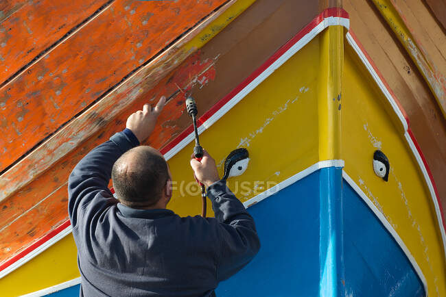 Vista trasera de un hombre pelando pintura de un barco luzzu tradicional con un soplete, Marsaxlokk, Malta - foto de stock