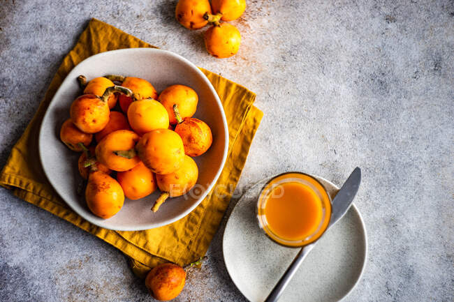 Top view of bowl of loquats on cloth napkin and plate with juice glass and knife on concrete surface — Stock Photo