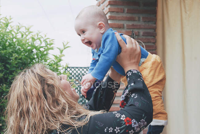 Femme souriante debout dans le jardin tenant son bébé garçon dans les airs — Photo de stock
