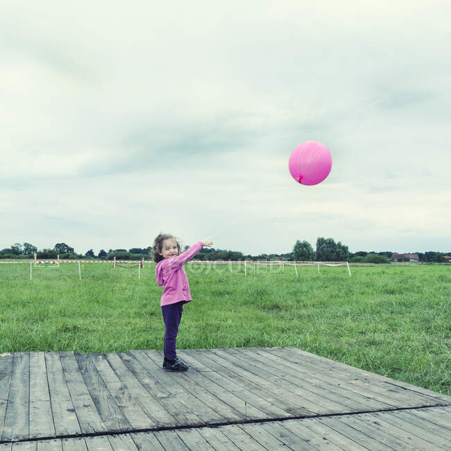 Chica sonriente de pie en un prado sosteniendo un globo rosa, Polonia - foto de stock