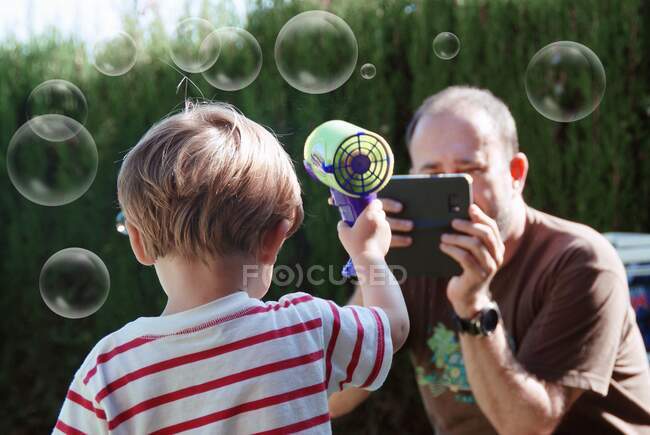 Hombre fotografiando a su hijo jugando con una pistola de jabón en el jardín - foto de stock