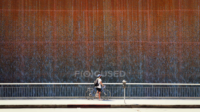 Teenage boy standing with his bicycle on pavement by a waterfall on a wall, USA — Stock Photo
