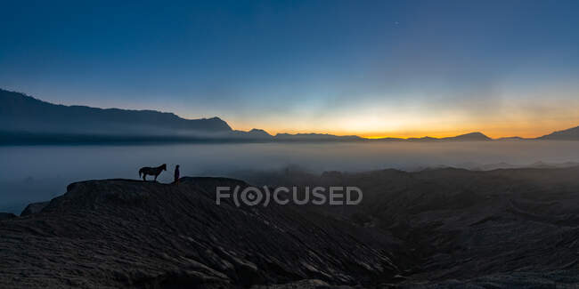 Silhueta de homem e seu cavalo ao pôr do sol no fundo das montanhas — Fotografia de Stock