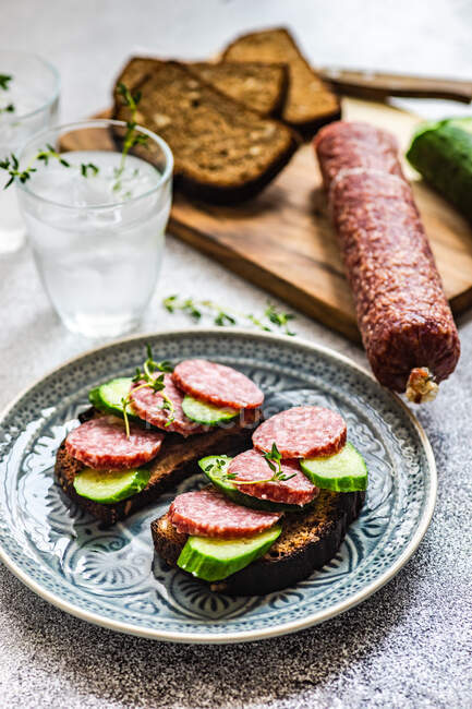 Slices of salami, cucumber and rye bread on a  chopping board with two glasses of water — Stock Photo