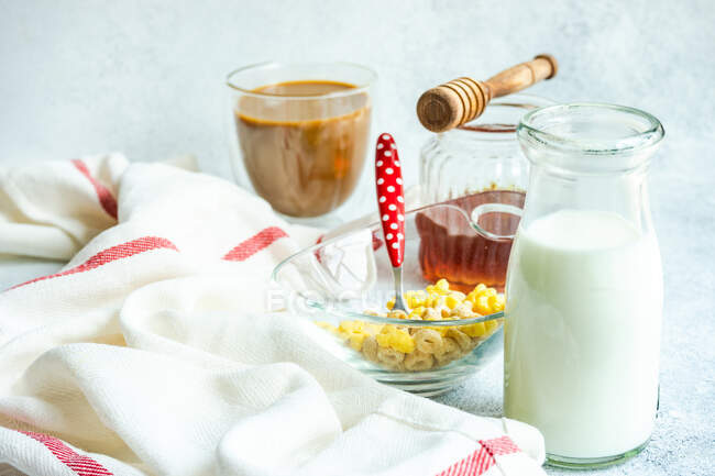 Bowl of cereal, milk, honey and a cup of coffee on a table next to a tea towel — Stock Photo