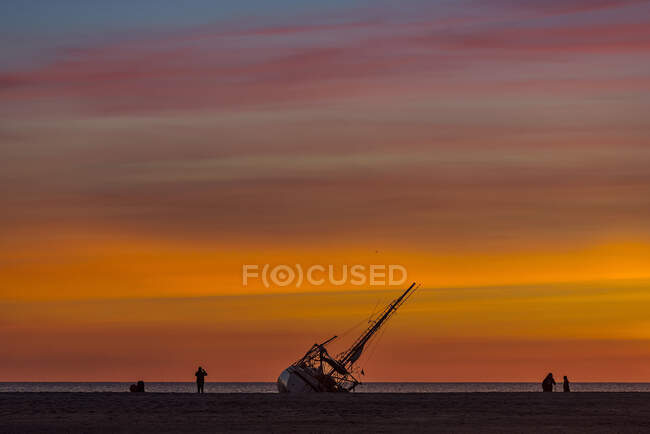 Silueta de tres personas en la playa junto a un naufragio al atardecer, Playa de Los Lances, Tarifa, Provincia de Cádiz, Andalucía, España - foto de stock