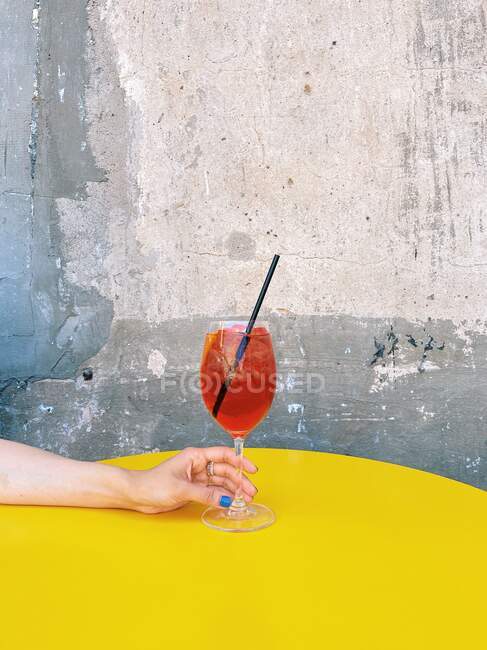 Женщина тянется за коктейлем из аперола на желтый стол — стоковое фото