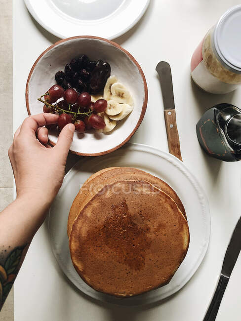 Person picking grapes from bowl with pancakes plate on table — Stock Photo