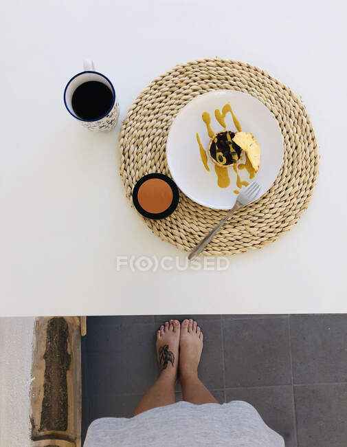 Pov shot of female feet next to table with cup of coffee and vegan chocolate, pineapple cake — Stock Photo