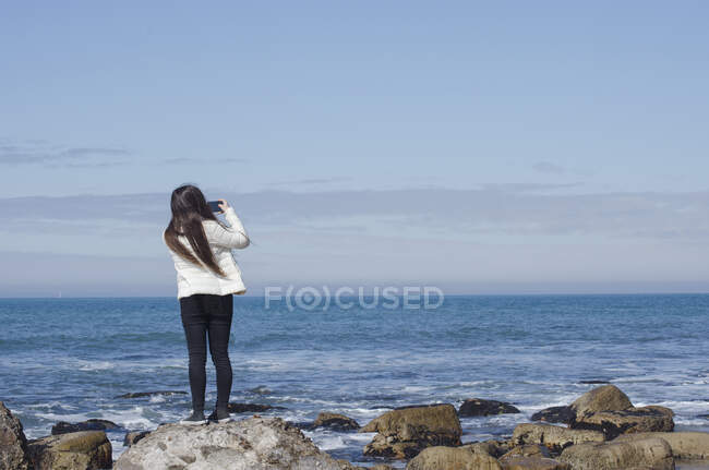 Rear view of a teenage girl standing on a rock taking a photo, Mar del Plata, Buenos Aires Province, Argentina — Stock Photo