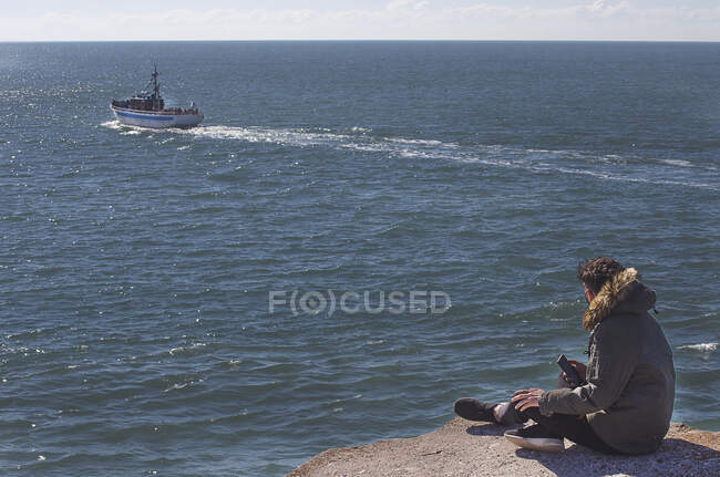 Человек, сидящий на скале у океана и смотрящий на плывущую мимо лодку, Мар-дель-Плата, провинция Буэнос-Айрес, Аргентина — стоковое фото