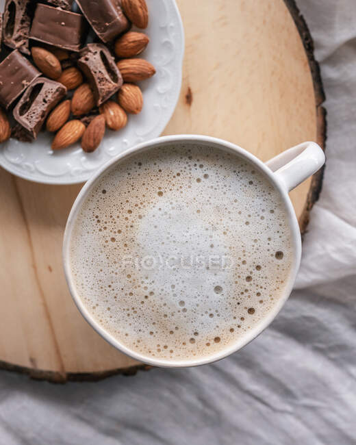 Cup of milky coffee next to plate with almonds and sliced chocolate bar — Stock Photo
