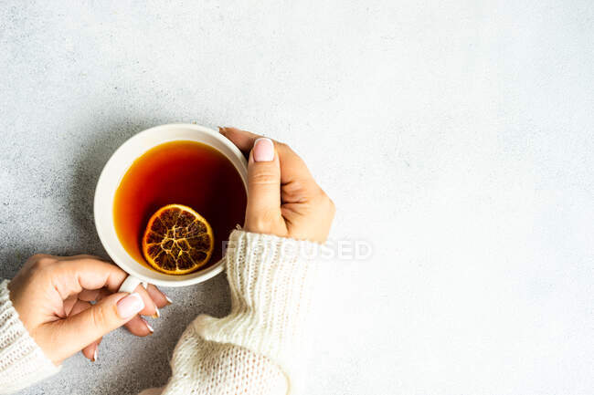 Вид зверху на жіночі руки, що тримають чашку з чаєм та сушеним апельсиновим шматочком — стокове фото