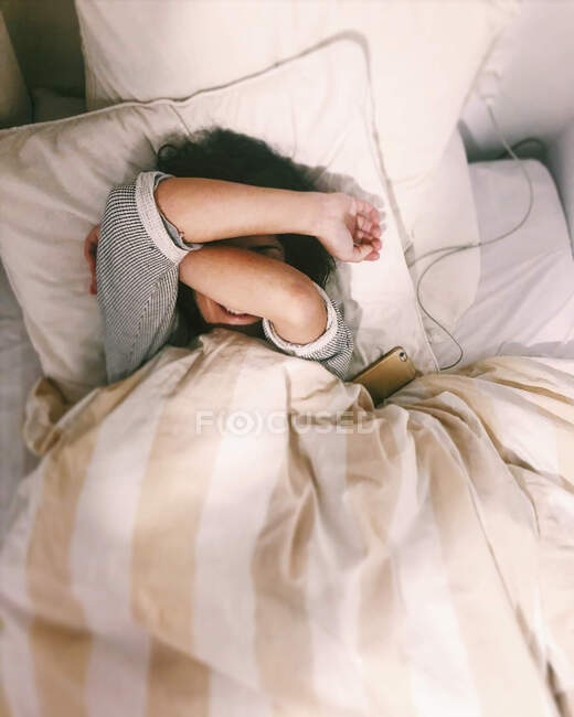 Smiling woman lying in bed next to her mobile phone with her arms covering her face — Stock Photo