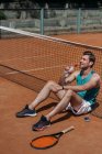 Молодой счастливый человек сидит на земле и пьет воду после теннисных тренировок — стоковое фото