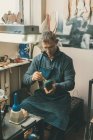 Zapatero maduro sosteniendo la pieza de trabajo de la bota y trabajando con la suela mientras está sentado en su taller tradicional - foto de stock