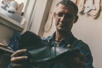 Selective focus of smiling mature cobbler in eyeglasses holding leather shoe in workshop — Stock Photo