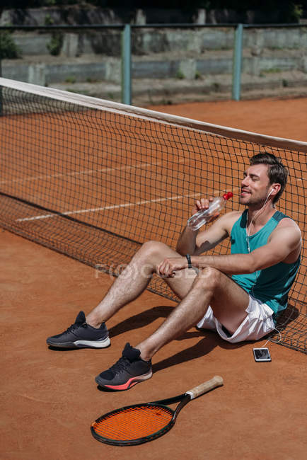 Joven feliz hombre sentado en el suelo y beber agua después del entrenamiento de tenis - foto de stock