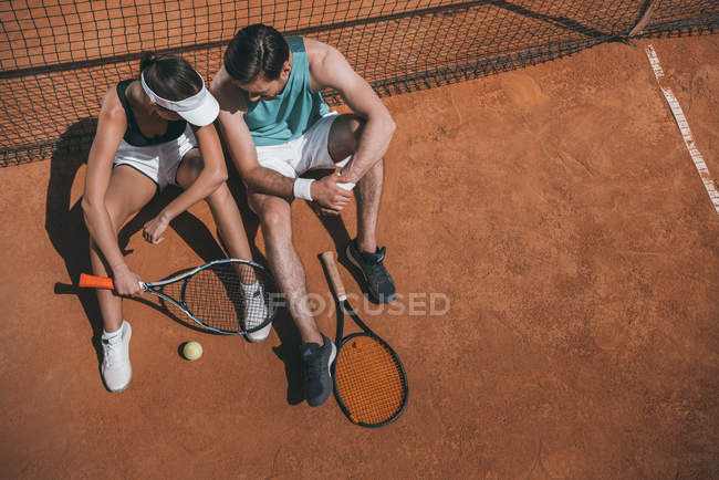 Vista de ángulo alto de la joven pareja apoyada en la red y relajante en la cancha de tenis - foto de stock
