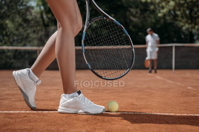 Ritagliato colpo di donna con racchetta in piedi sopra la palla da tennis sdraiato sul campo — Foto stock