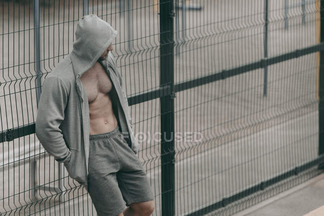 Sportif debout dans un sweat à capuche déboutonné près de la clôture sur le terrain de sport — Photo de stock