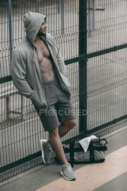 Sportivo in piedi in felpa con cappuccio sbottonato vicino alla recinzione al campo sportivo — Foto stock