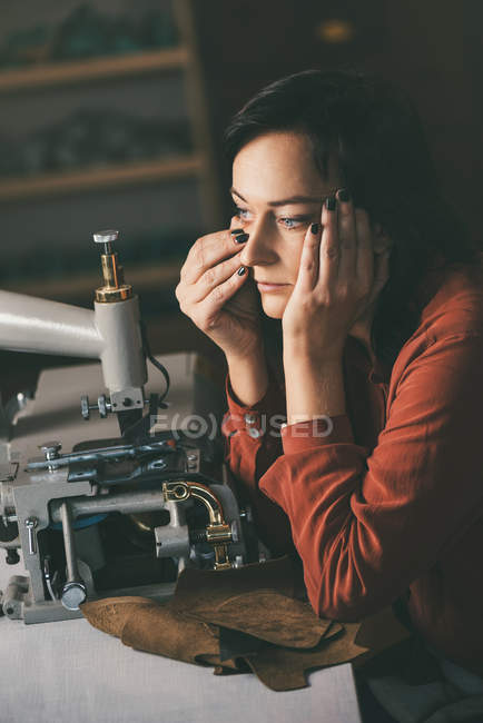 Задумчивая швея смотрит в сторону, работая с швейной машинкой и кожей в мастерской сапожника — стоковое фото
