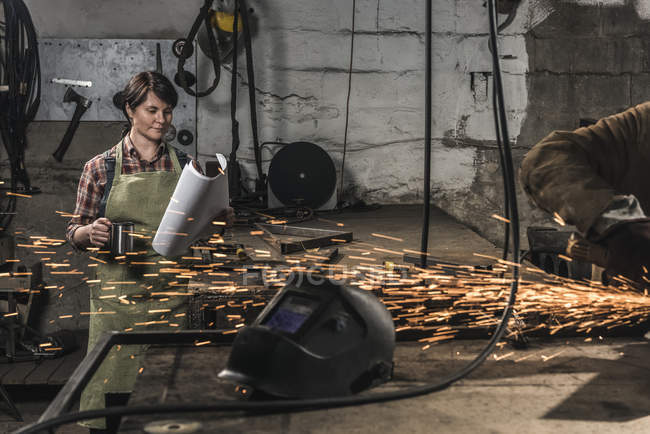 Welder with cup of hot drink reading papers while colleague working in workshop — Stock Photo