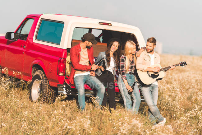 Group of happy young people drinking beer and playing guitar while relaxing in car trunk in flower field — Stock Photo