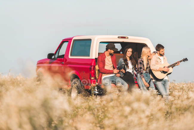 Group of young friends drinking beer and playing guitar while sitting in car trunk in flower field — Stock Photo